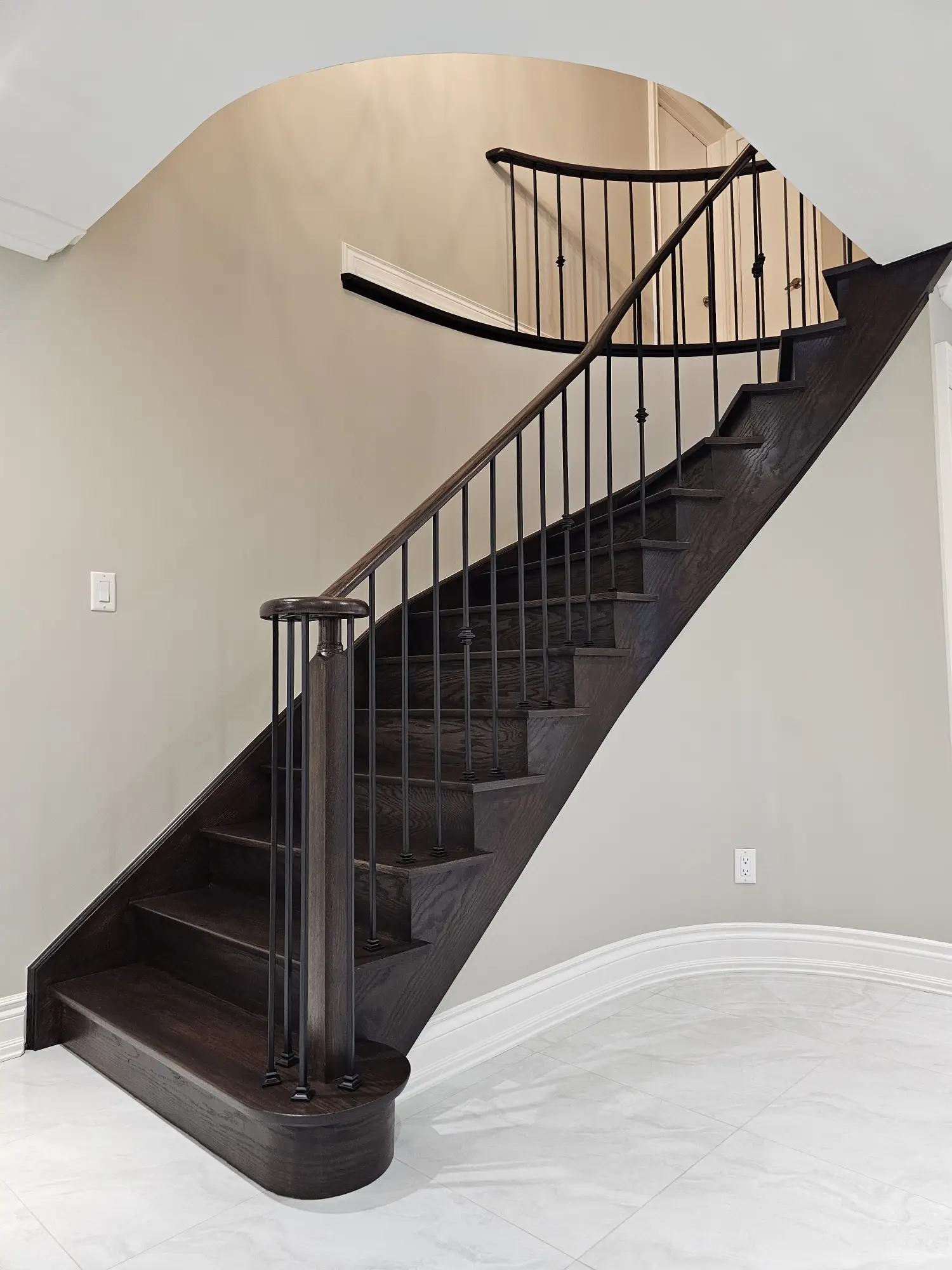 A curved staircase with polished dark wood steps, black balusters, and neutral walls, set above a glossy tile floor.