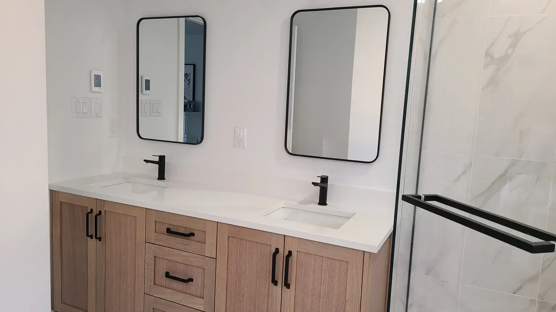 Oak double vanity bathroom renovation in Toronto with black faucets, black framed mirrors, quartz countertop, and glass shower beside the vanity.
