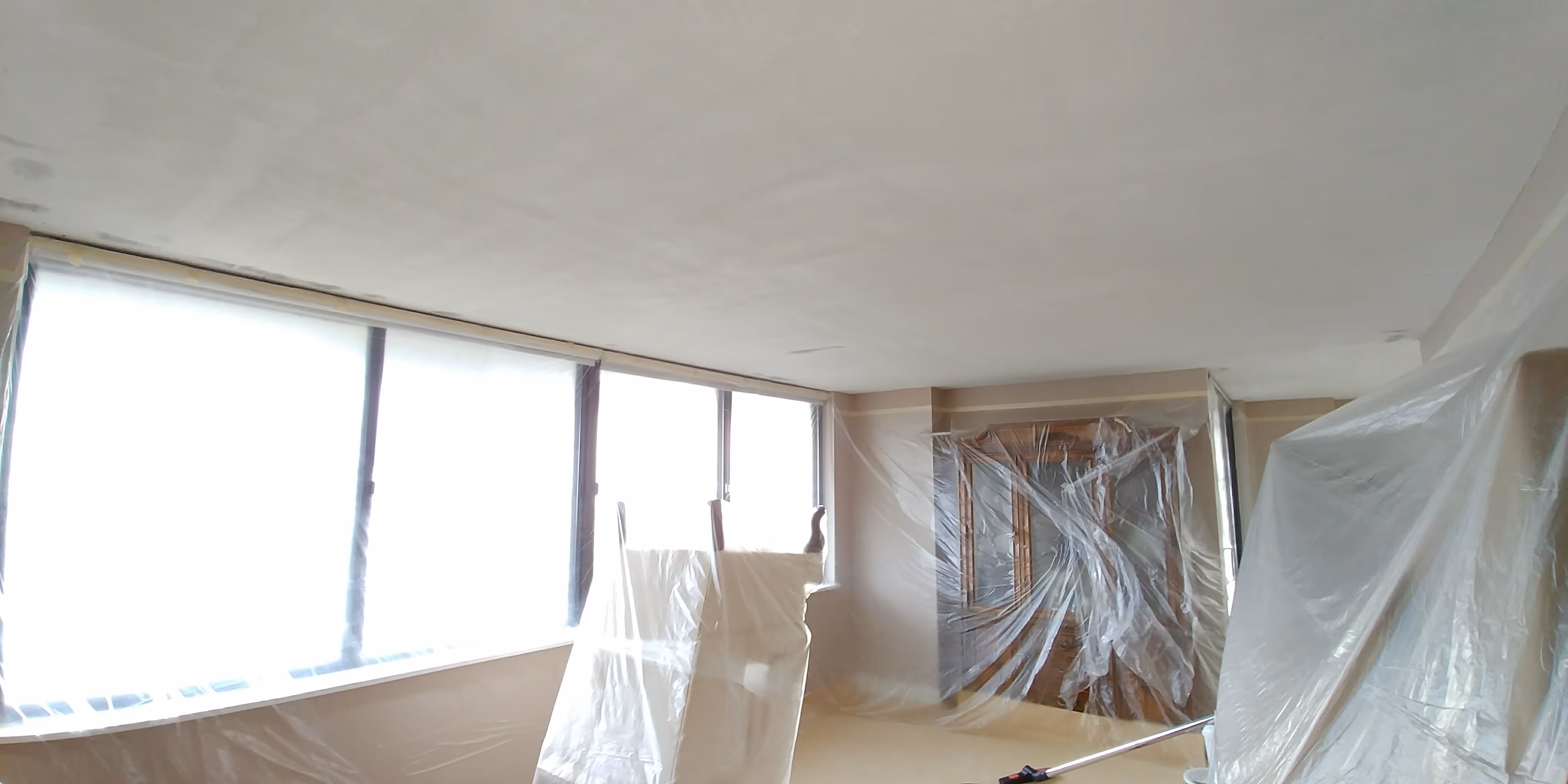 A freshly renovated room with smooth, dust-free ceilings, large windows letting in natural light, and furniture carefully covered for protection during the process.