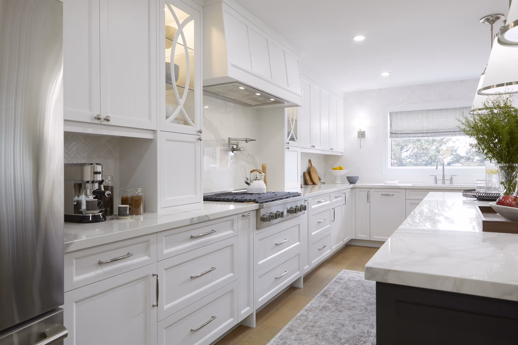 A bright, modern kitchen with sleek white cabinetry, marble countertops, stainless steel appliances, and a large window bringing in natural light.